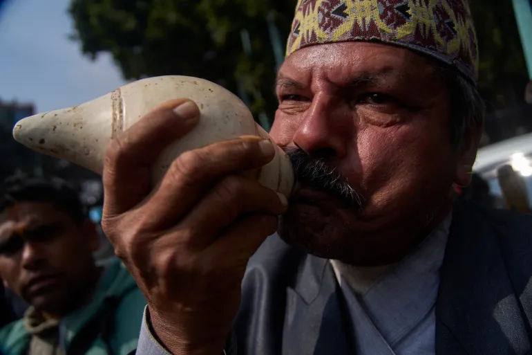 शुक्रवार, 13 फरवरी, 2026 को काठमांडू, नेपाल के त्रिभुवन अंतर्राष्ट्रीय हवाई अड्डे पर नेपाल के पूर्व राजा ज्ञानेंद्र शाह के आगमन पर उनके स्वागत के लिए इकट्ठा हुए लोगों के बीच एक समर्थक ने शंख बजाया। (एपी फोटो/निरंजन श्रेष्ठ)