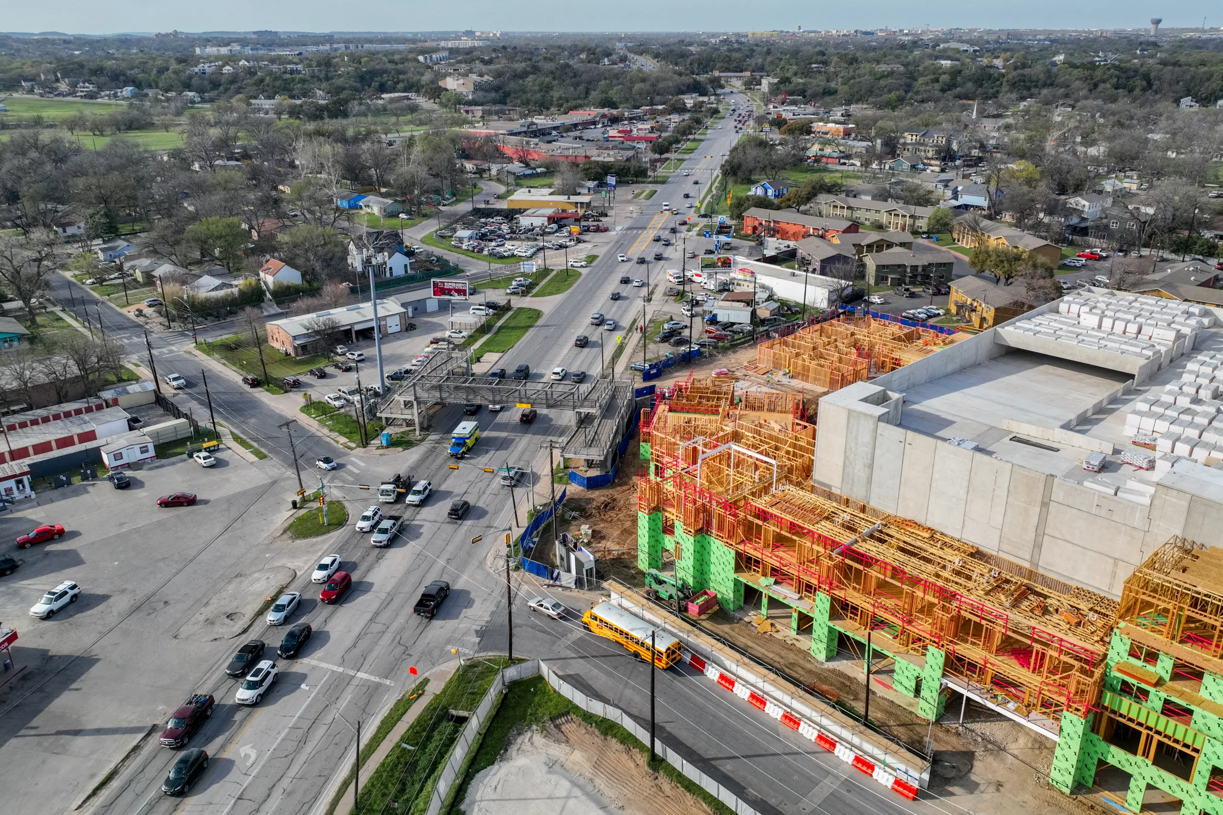 Aerial view of a multi-story apartment building under construction beside a busy intersection in Austin, Texas, surrounded by low-rise homes, businesses, and tree-covered neighborhoods.